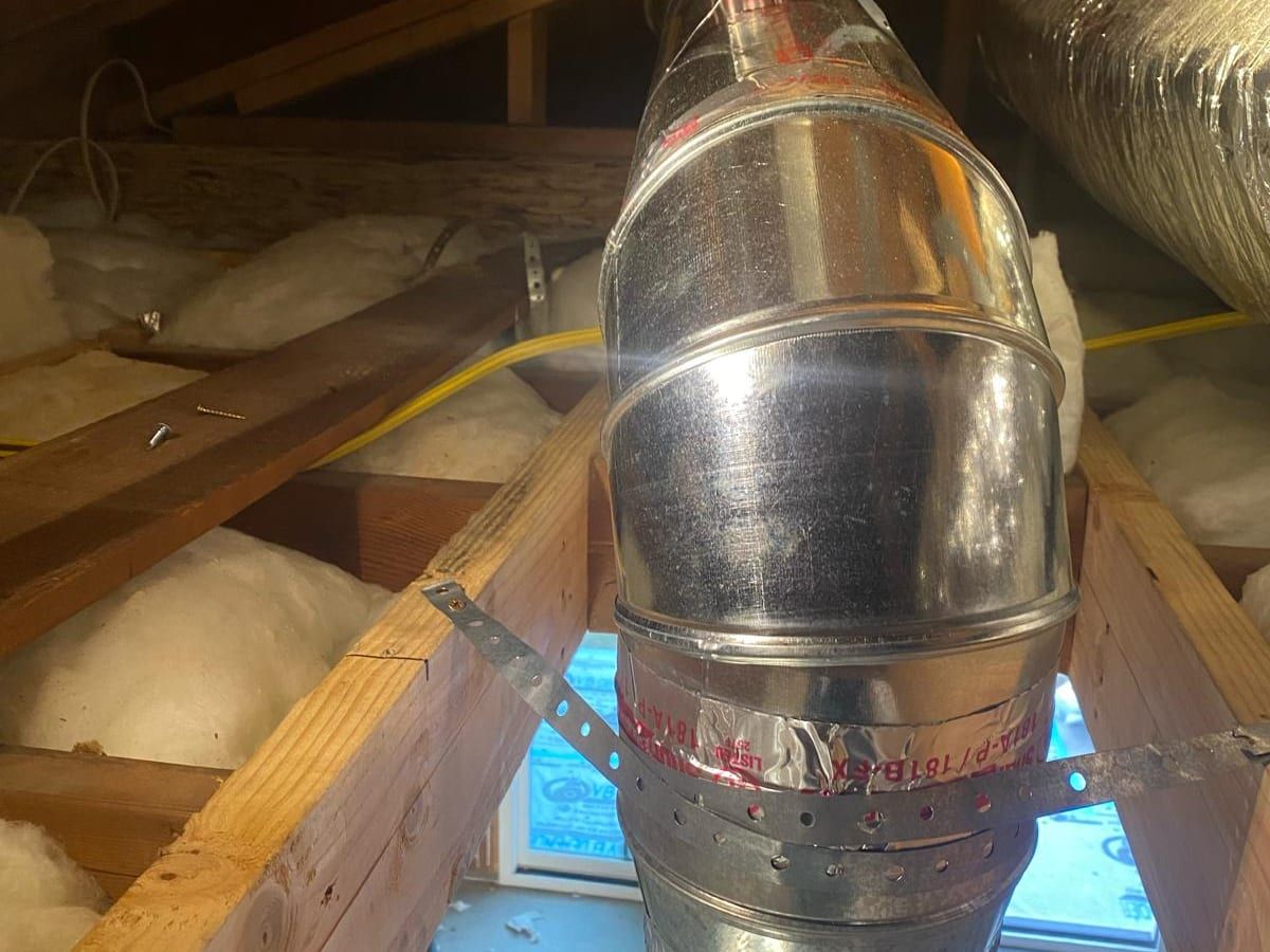 A metal vent pipe rising between attic rafters, held by a metal strap, with insulation and a flexible duct around it