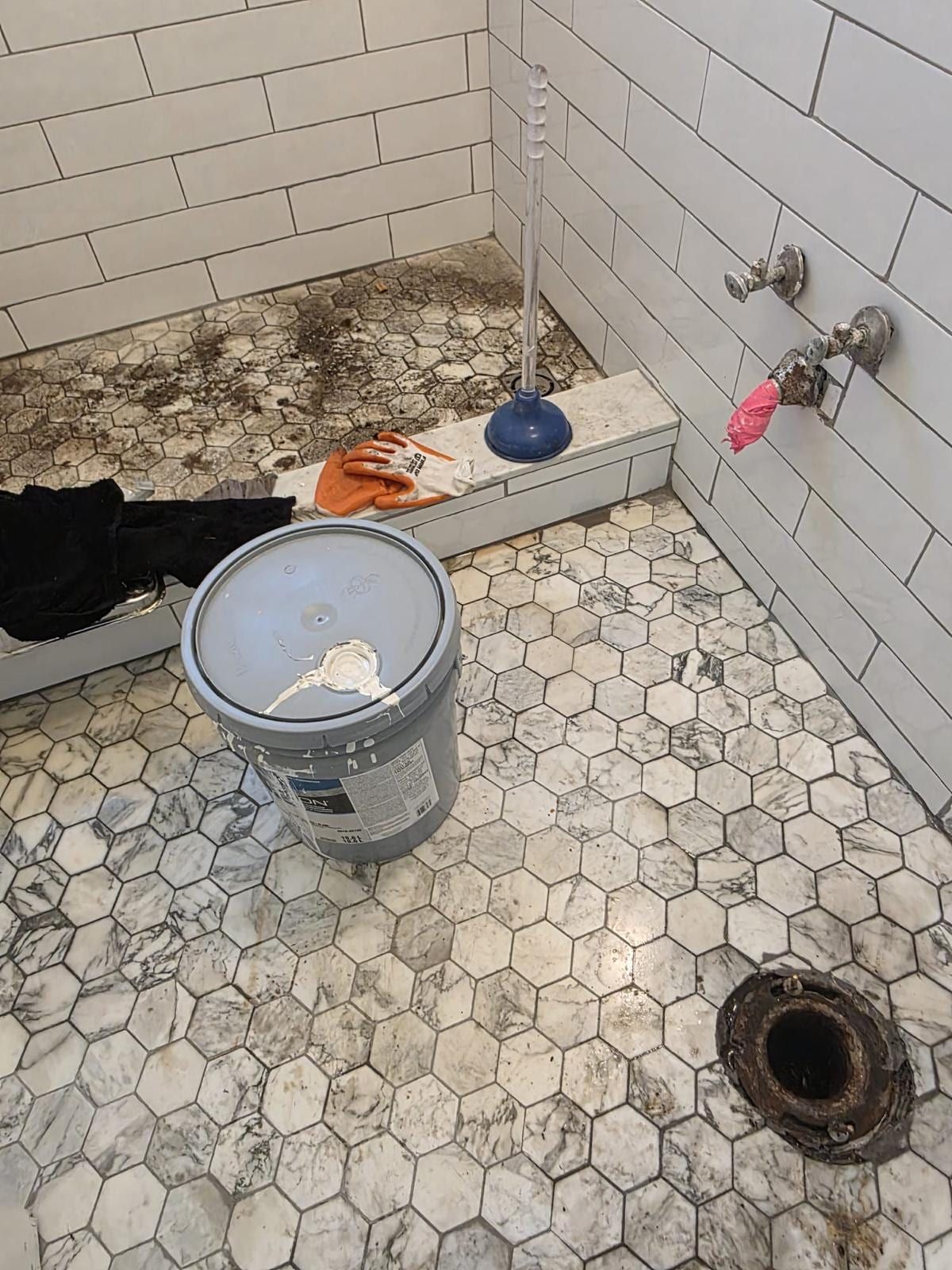 A bathroom being rebuilt: white hexagon floor tile around an open toilet flange, white subway tile walls with two valve stubs, a bucket, a plunger and work gloves