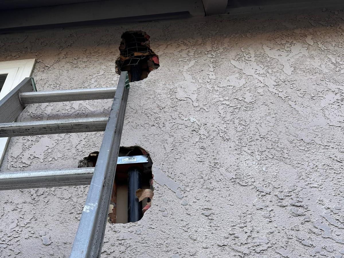 Two openings cut in a stucco exterior wall beside an aluminum ladder, with pipe visible inside the wall